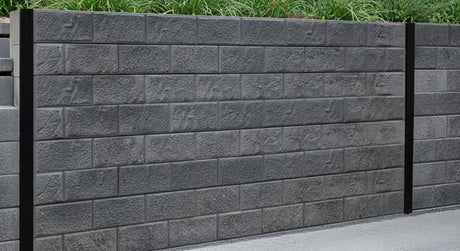 Modern backyard retaining wall using Smooth Charcoal Concrete Sleepers with black steel H-posts, bordering a white pebble driveway and leveled green lawn.