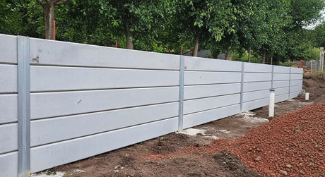 Long residential retaining wall installation using smooth light grey concrete sleepers and galvanised steel I-beam posts, bordering a garden bed with red volcanic mulch.