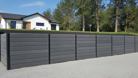 Modern residential backyard featuring a sleek Charcoal Smooth Concrete Sleeper retaining wall installed with black steel posts, bordering a white pebble driveway and green lawn.