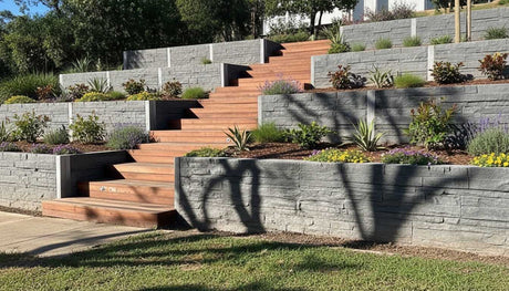 Professional installation of a multi-level terraced retaining wall using Stacked Stone Charcoal Concrete Sleepers, featuring integrated timber garden stairs and drought-tolerant landscaping.