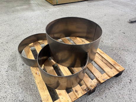 Three various-sized industrial round metal planter or fire pit liners displayed on a wooden pallet in a warehouse.